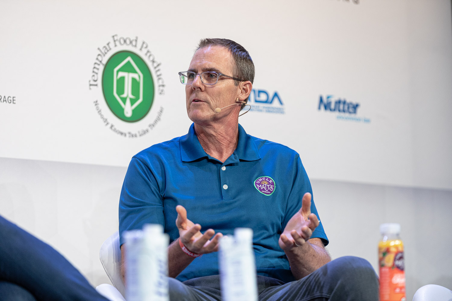 A man with glasses in a blue polo with an Uncle Matt's logo holds out both hands, with juice bottles on the table in front of him.