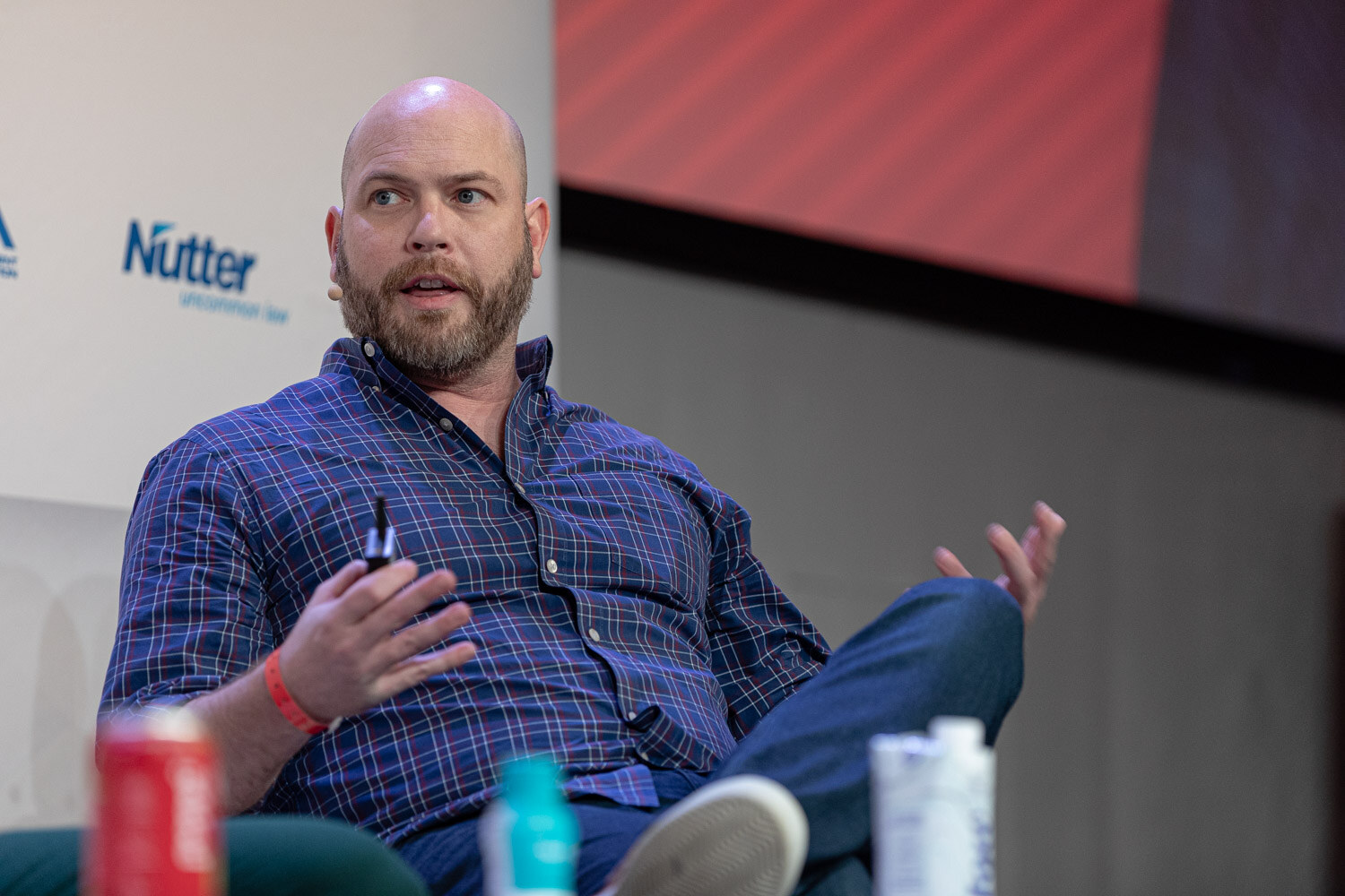 A bearded man in a navy plaid shirt sits with an open palm raised, a Coca-Cola style can and bottle in front of him.