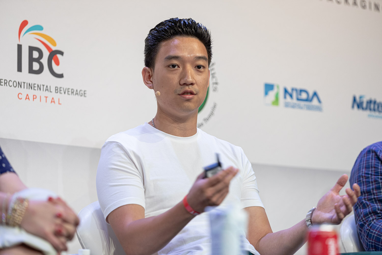 A man in a white crew-neck t-shirt holds a small device and gestures with his open hand in front of IBC and NIDA logos.