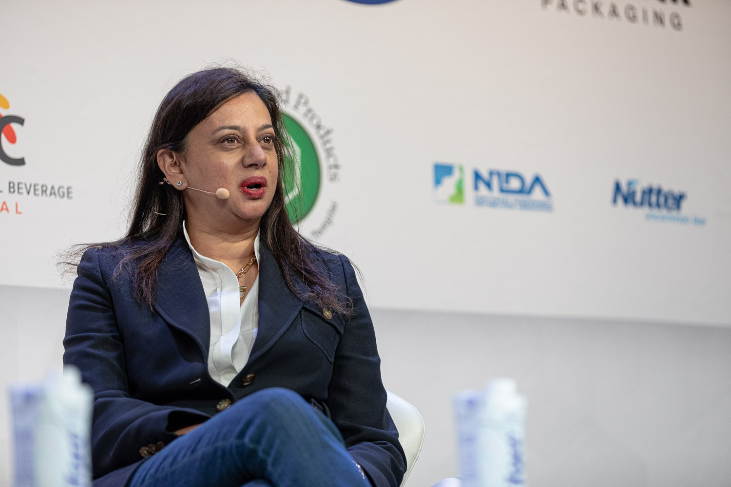 Close view of a woman with dark hair in a navy blazer and white blouse speaking, with sponsor logos behind her.