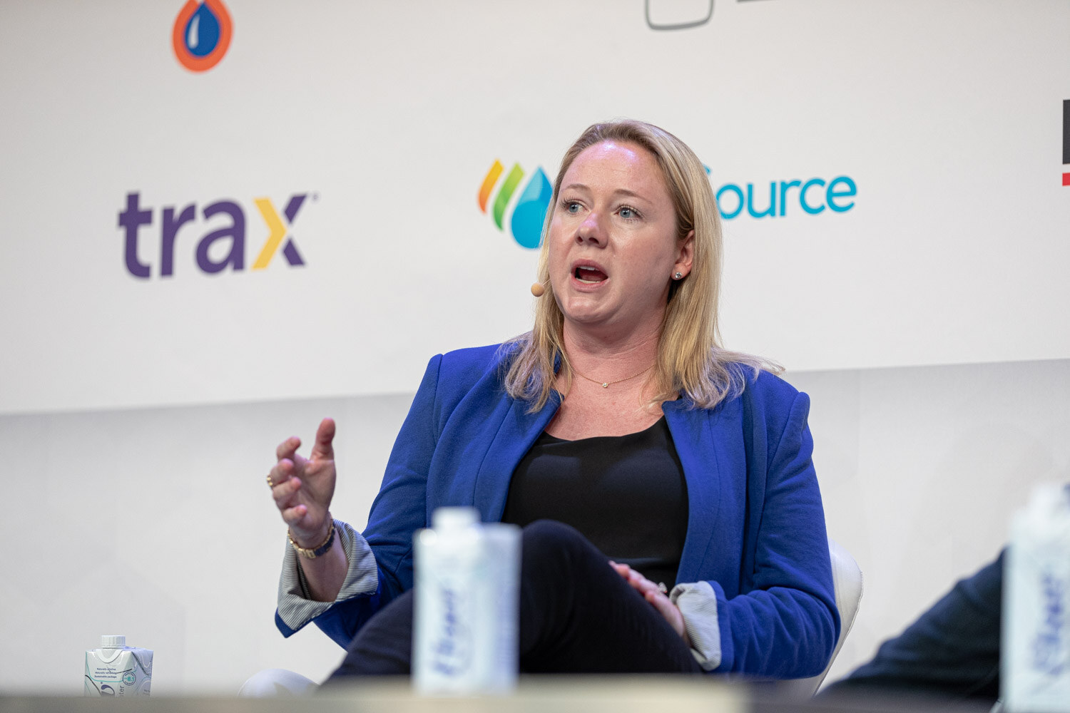 A woman in a blue blazer speaks into a headset microphone, her right hand raised, with Trax and BevSource signs behind her.