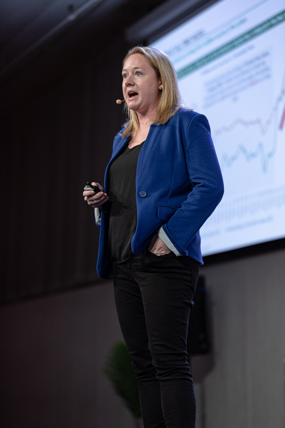 A woman in a blue blazer and black pants stands at the edge of the stage with a chart on the screen behind her.