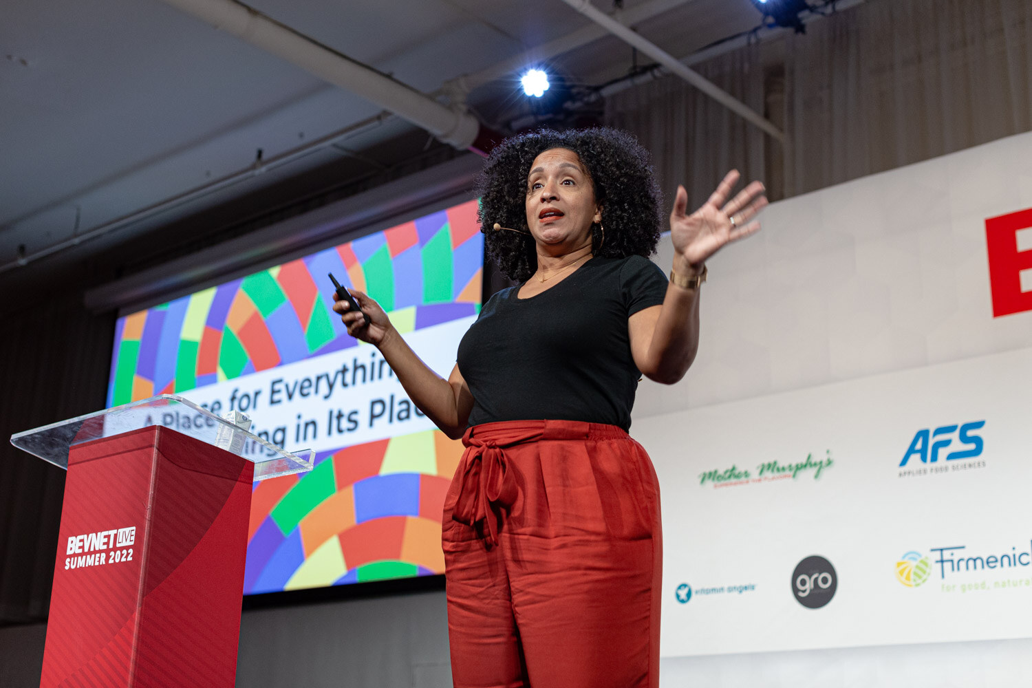 A woman in a black shirt and red pants gestures with a clicker in front of a screen with a multicolor pattern.