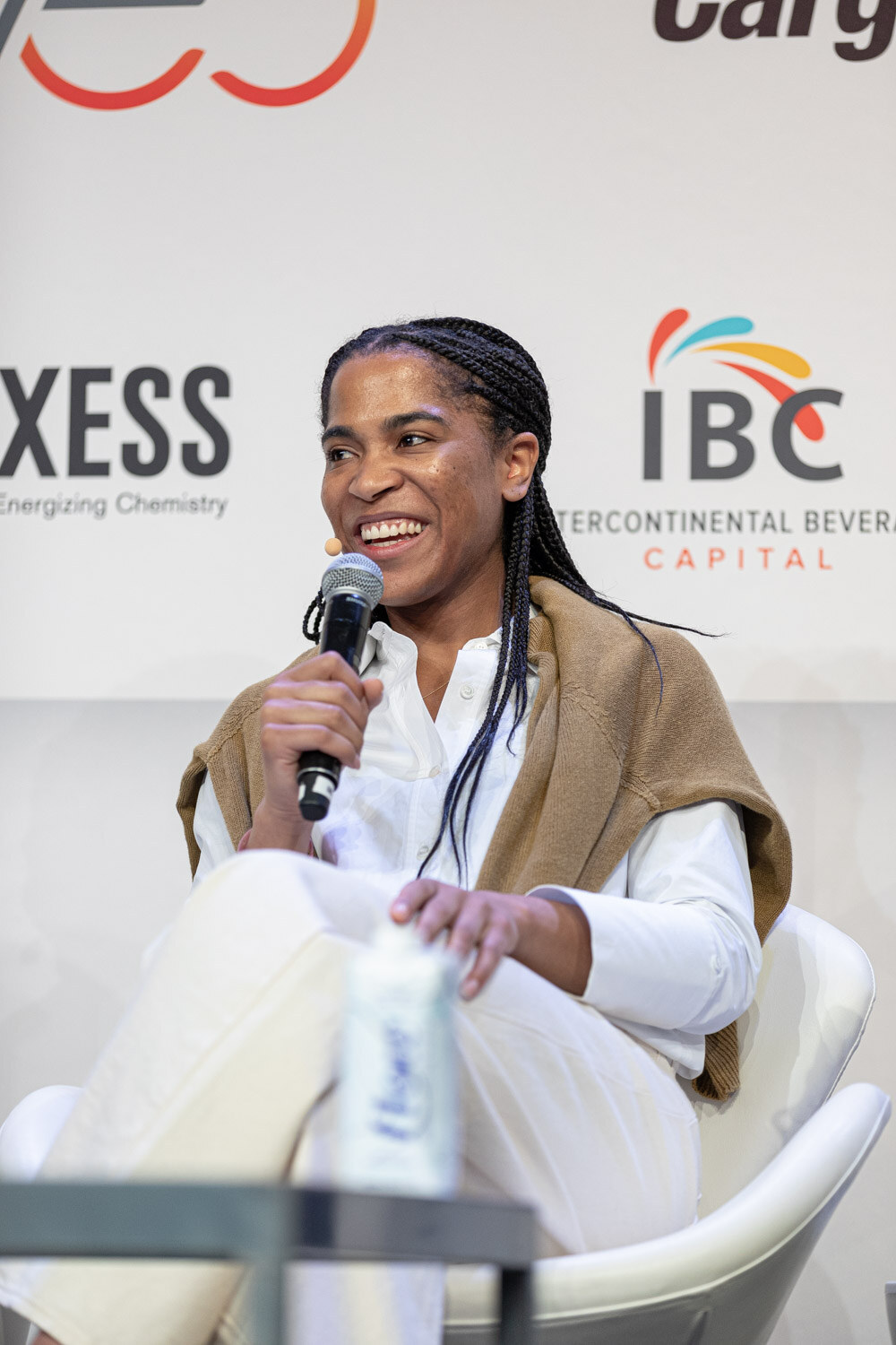 A woman with braids in a white shirt and tan sweater laughs, holding a microphone, in a white chair.