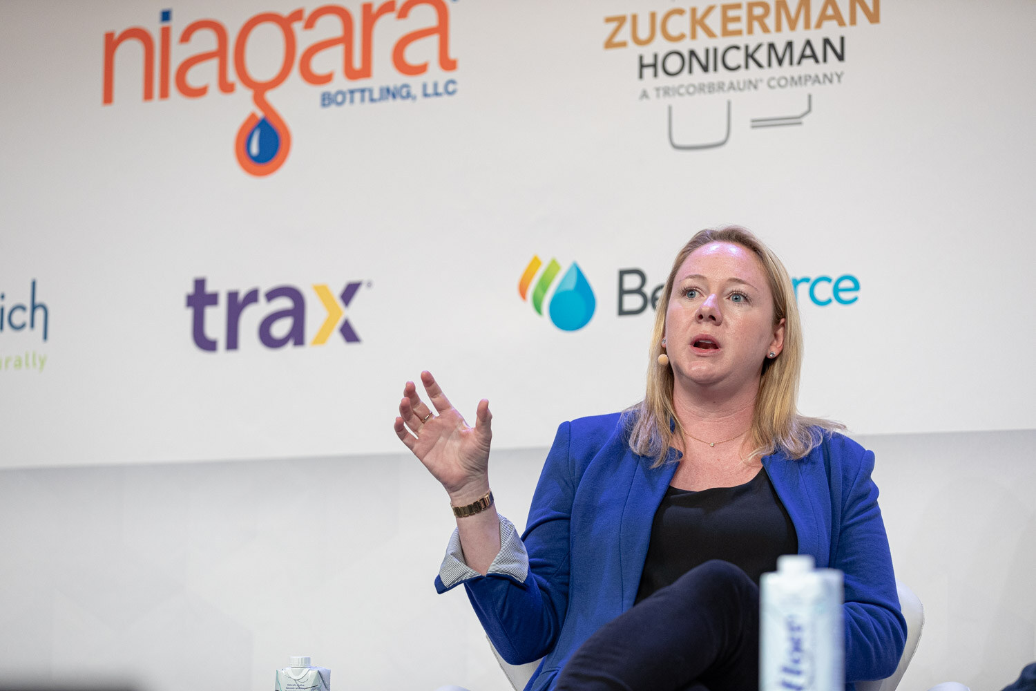 A woman with blonde hair in a blue blazer gestures with her left hand, with Trax and BevSource logos behind her.