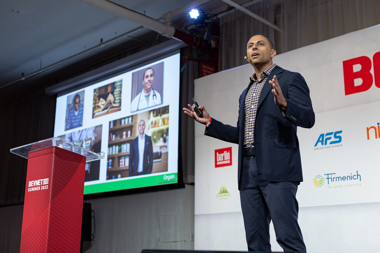 A man in a navy blazer stands by a red podium with a projector screen showing small photos of people and shelves of products.
