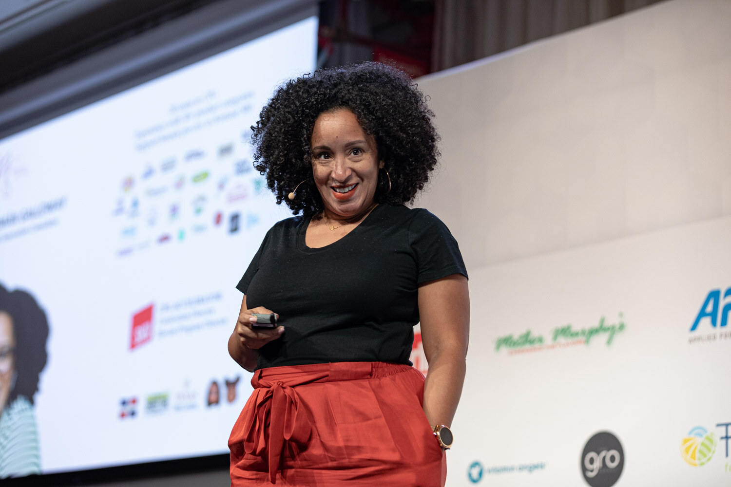 A woman with curly hair in a black shirt and red skirt holds a clicker and smiles on stage.