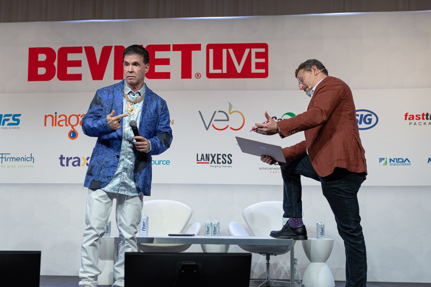 Two men on stage under a BevNET Live backdrop, one in a blue jacket pointing, the other in a rust jacket holding a laptop.