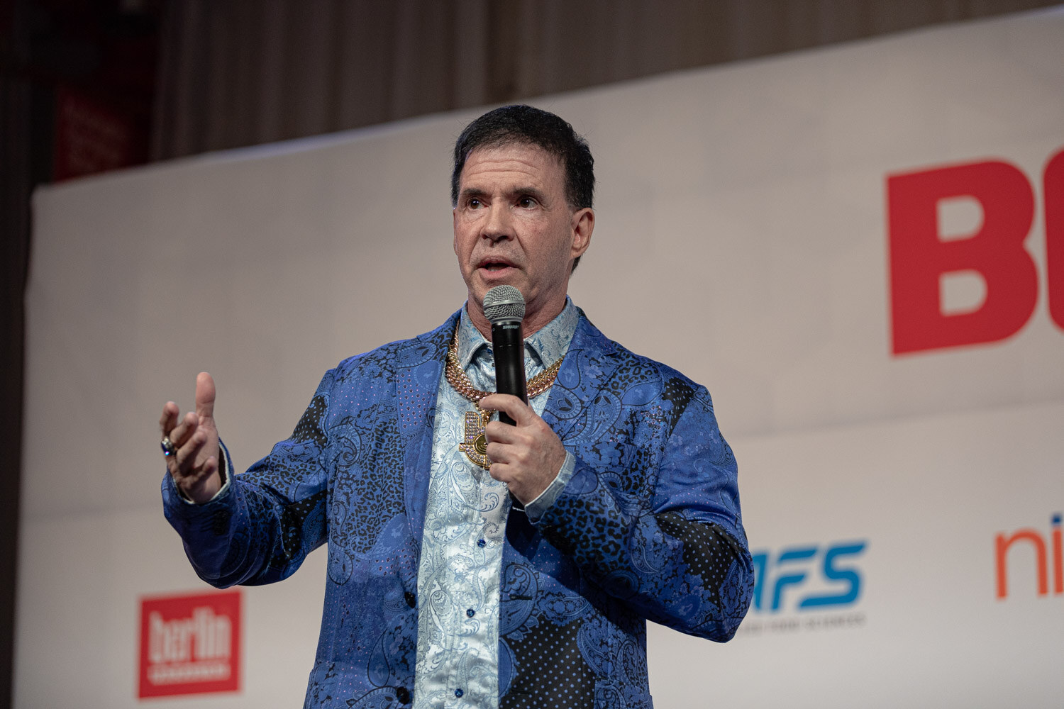 A man in a blue patterned jacket and gold chains speaks into a microphone on the BevNET Live stage.