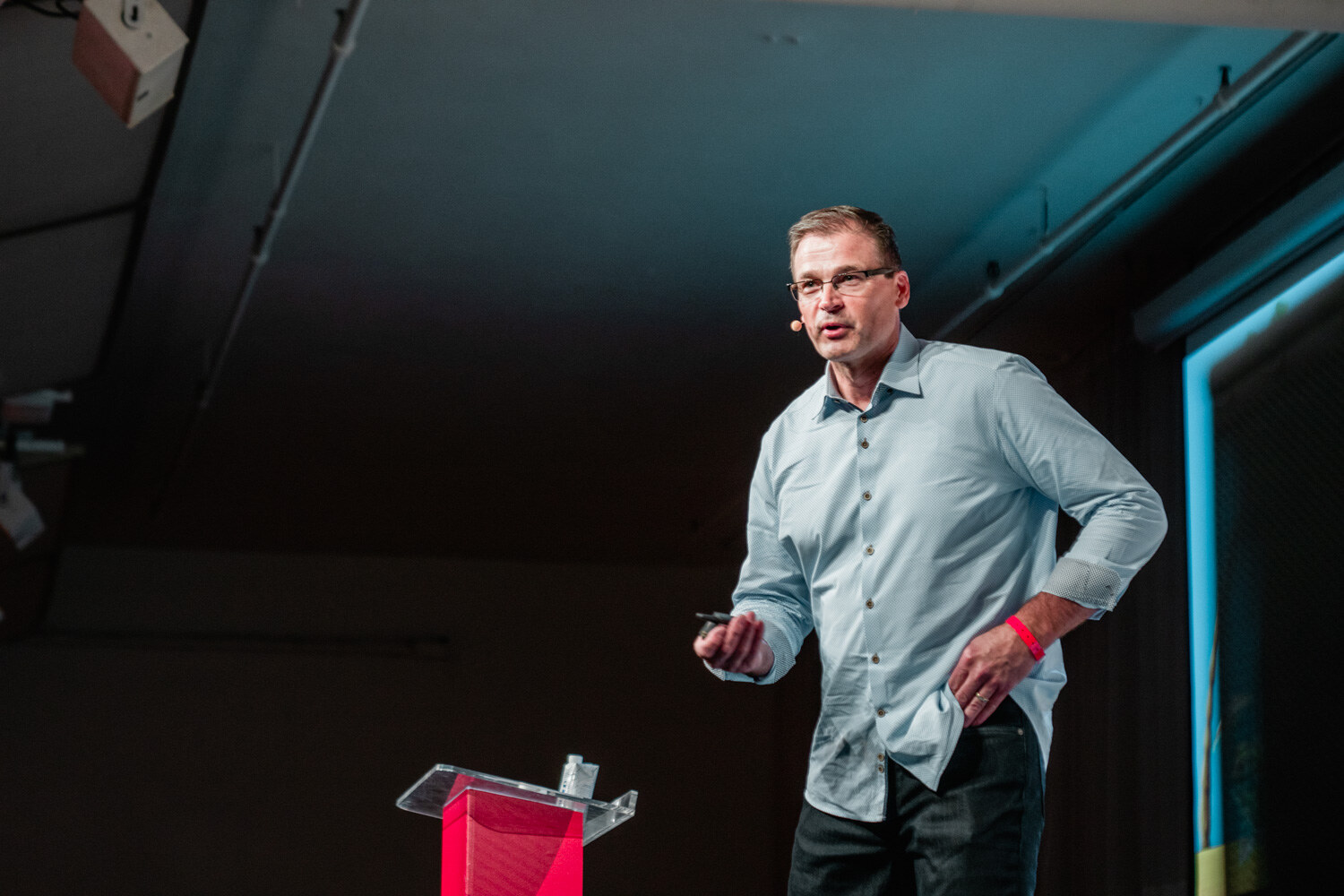 A man in a light blue shirt and glasses walks on a darkened stage beside a red podium.