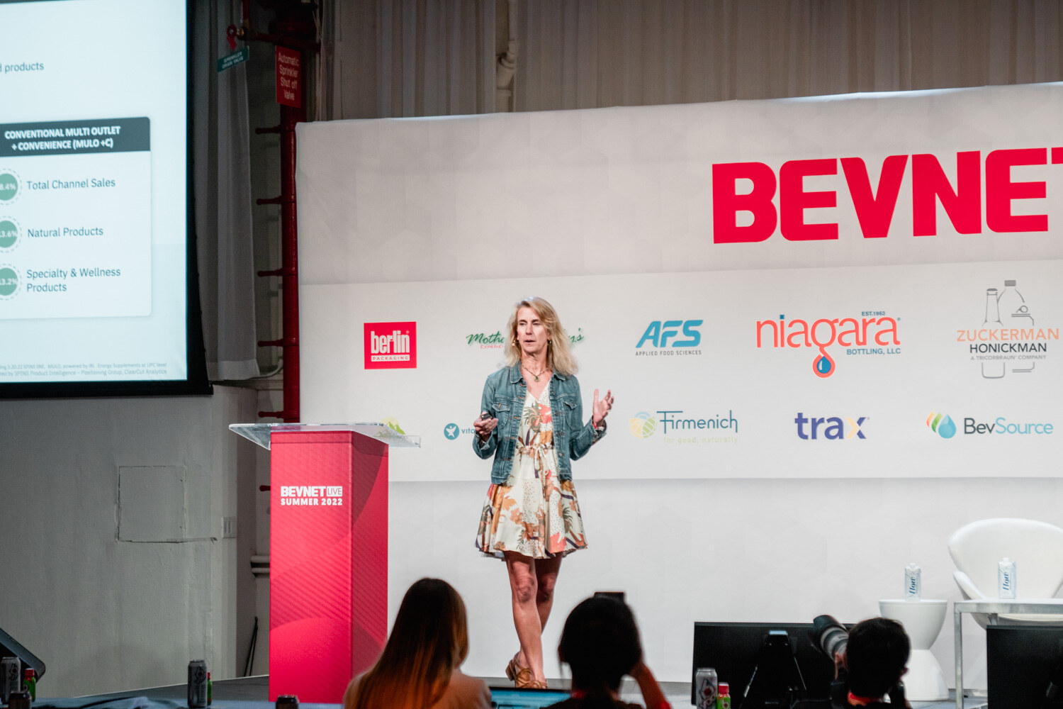 A woman in a denim jacket and floral dress stands in front of a BevNET Live backdrop with a large screen of charts at the left.