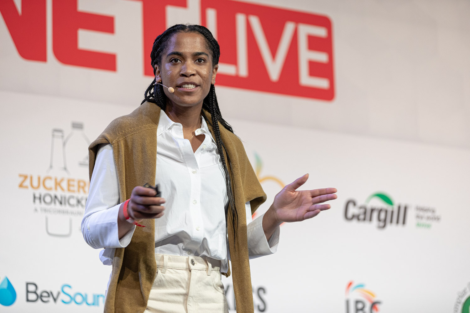 A woman in a white shirt and tan sweater tied at the shoulders gestures with a clicker, with sponsor logos behind her.