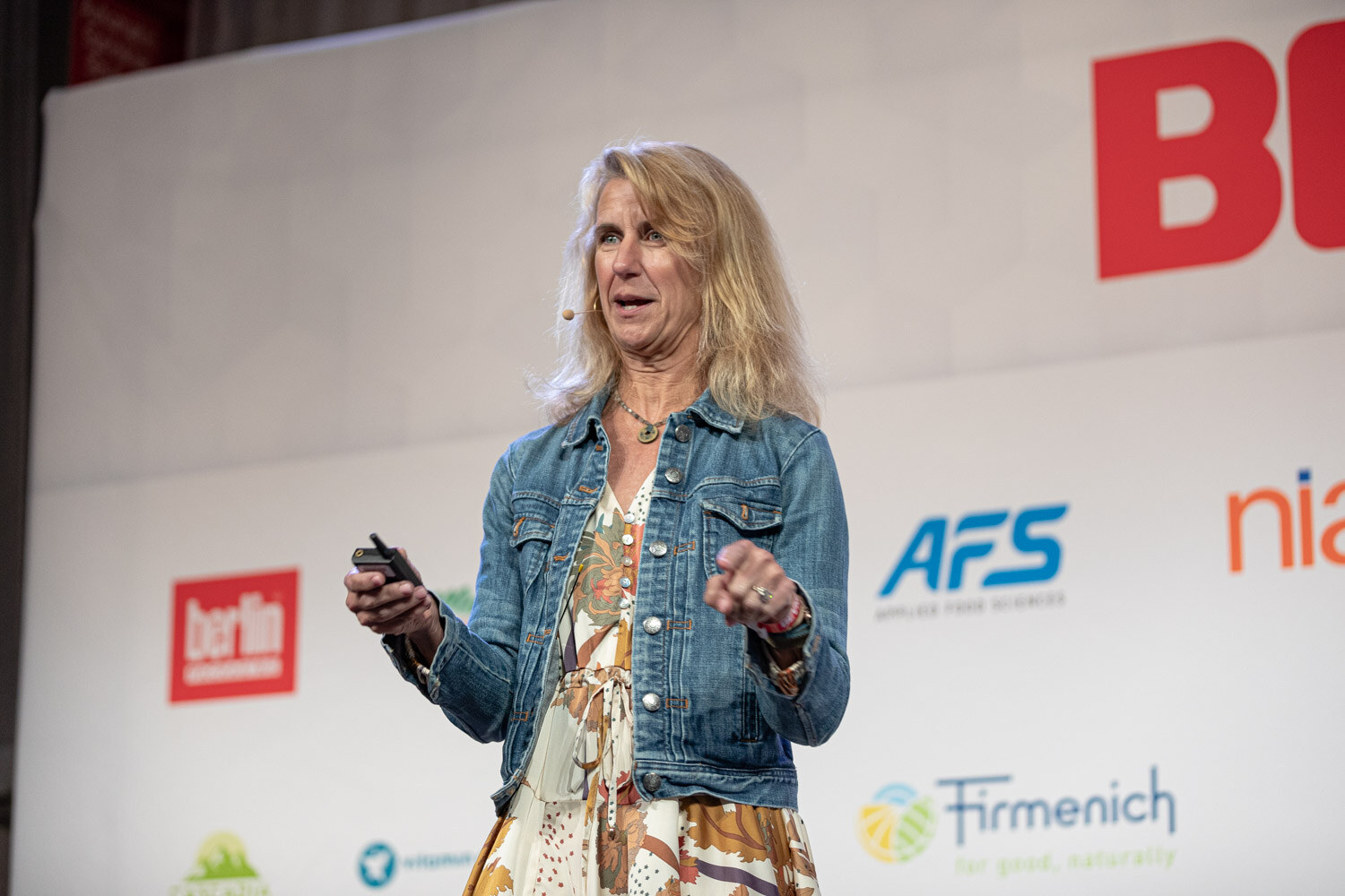 A woman with blonde hair in a denim jacket and floral dress holds a clicker in front of a BevNET Live backdrop.