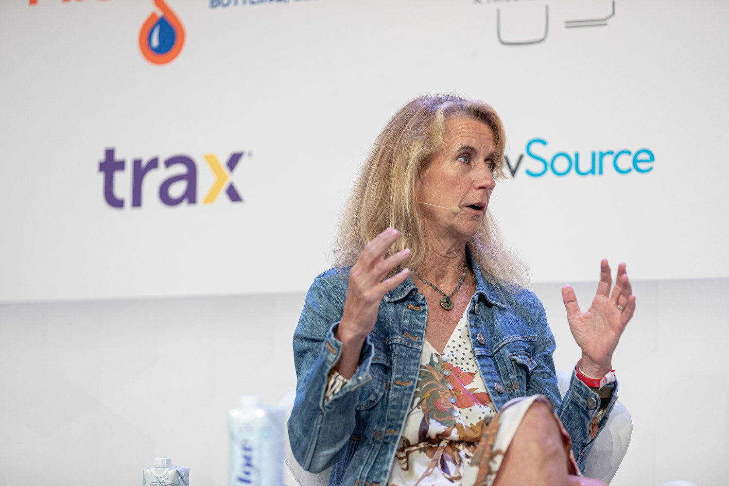 A woman with long blonde hair in a denim jacket and floral dress gestures with both hands while seated, in front of Trax and BevSource signs.
