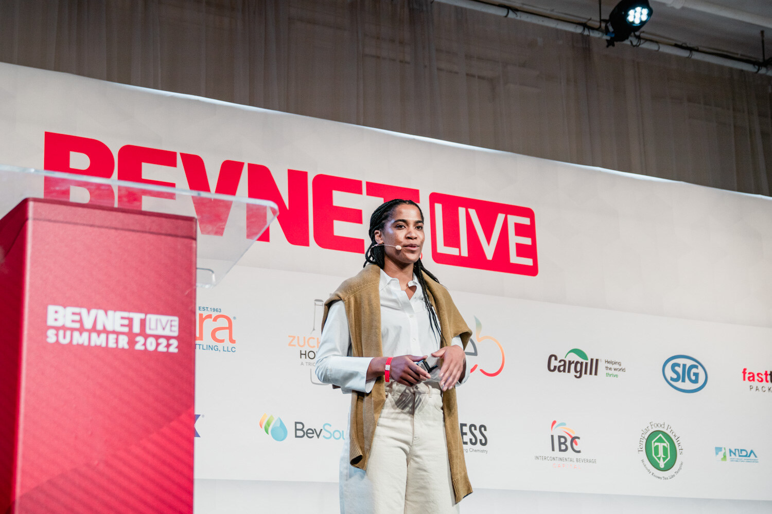 A woman in a tan sweater and white pants stands on stage beside a red podium reading BevNET Live Summer 2022.