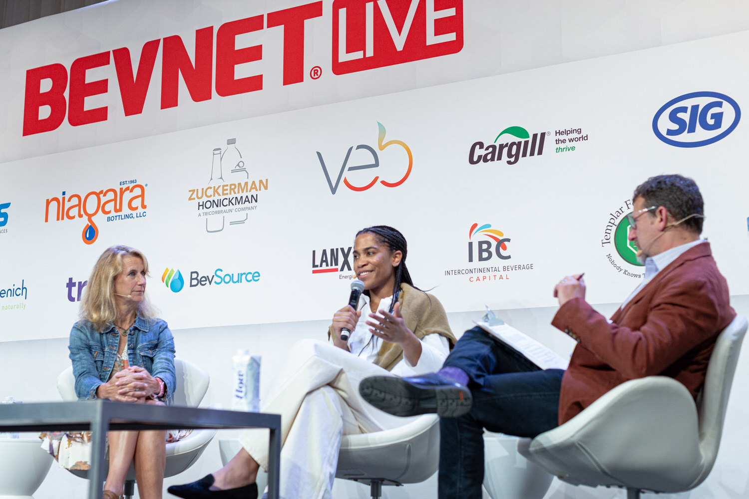 A woman with braids speaks with a microphone while a woman in a denim jacket and a man in a rust jacket sit in white chairs.
