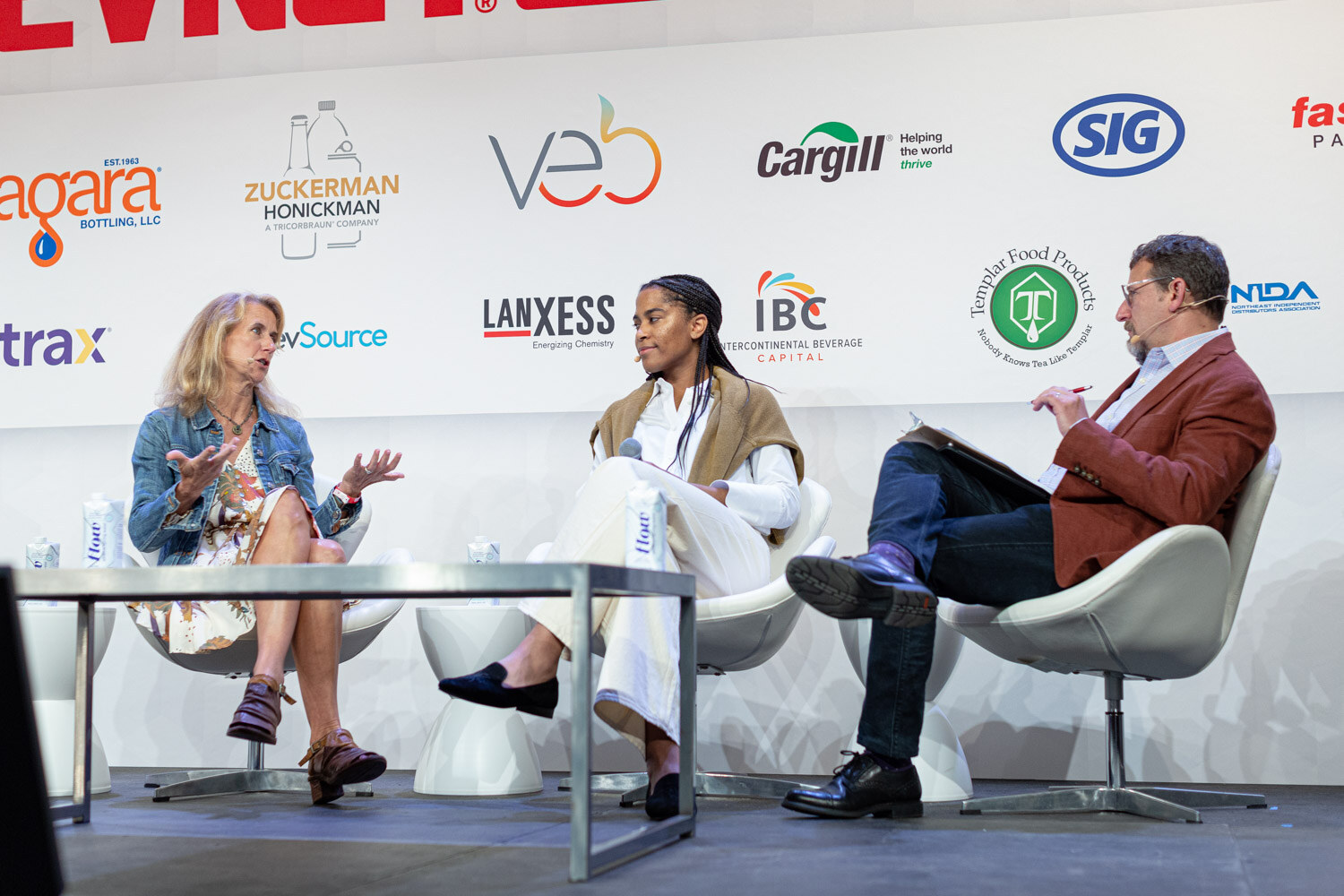 A woman in a denim jacket, a woman in white pants and a man in a rust jacket sit across a low table on stage.