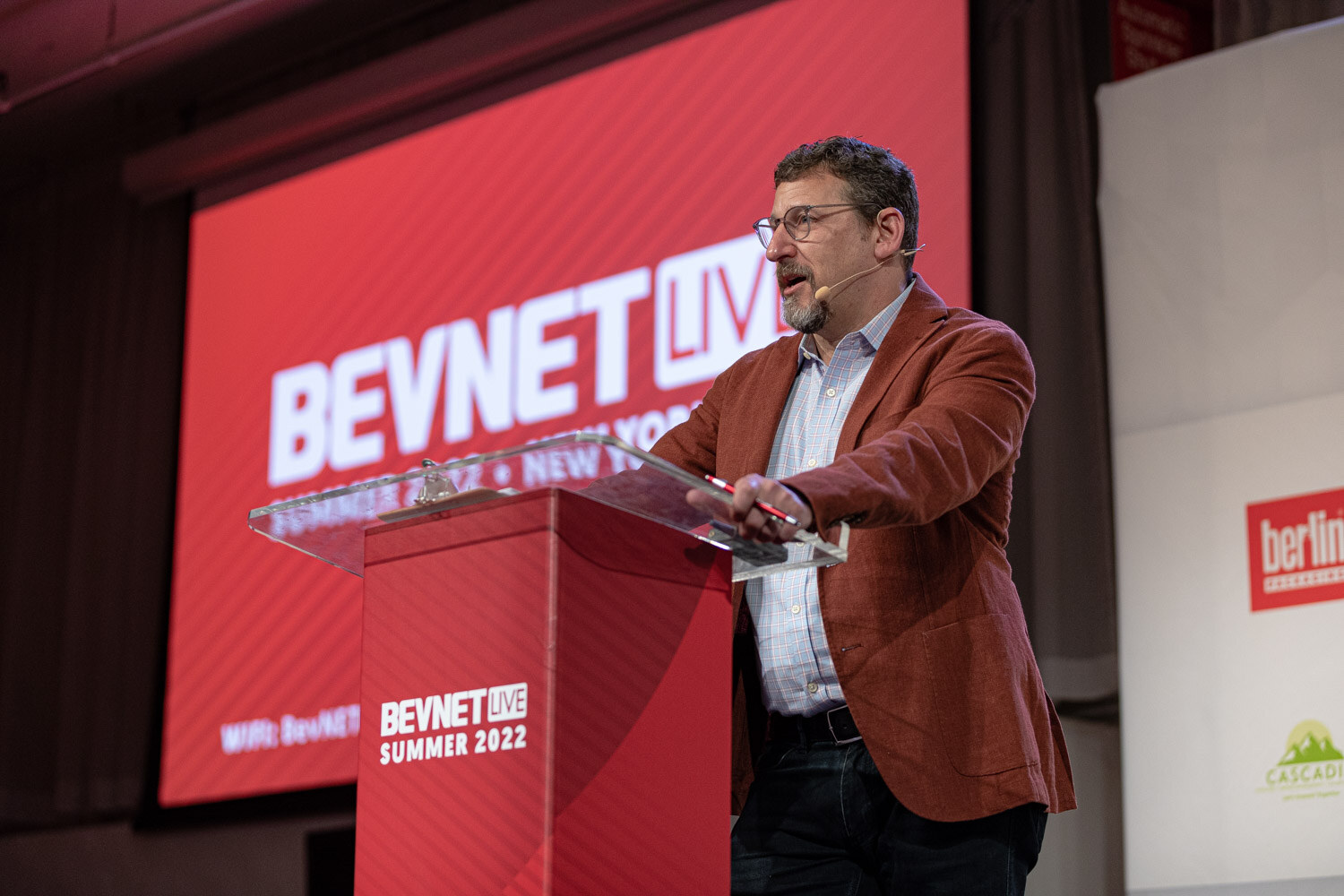 A man in a rust-colored jacket and glasses speaks from a clear podium in front of a red BevNET Live screen.
