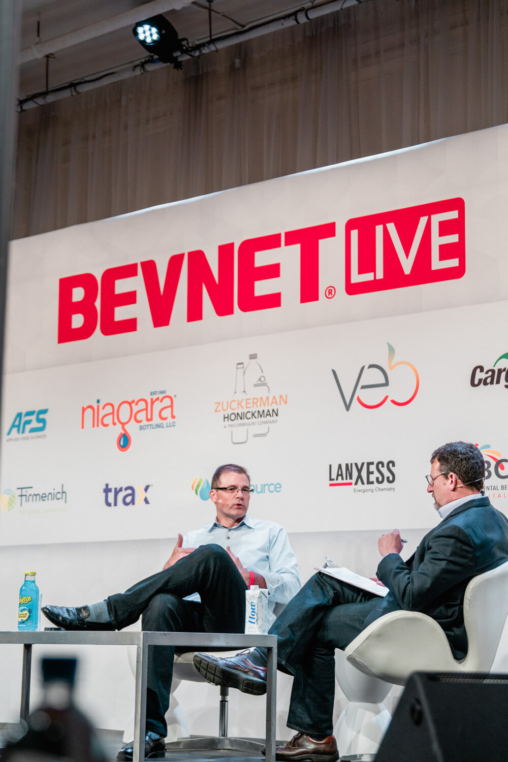 Two men sit in white chairs below the BevNET Live sign and sponsor logos, a bottle of lemonade on the table between them.