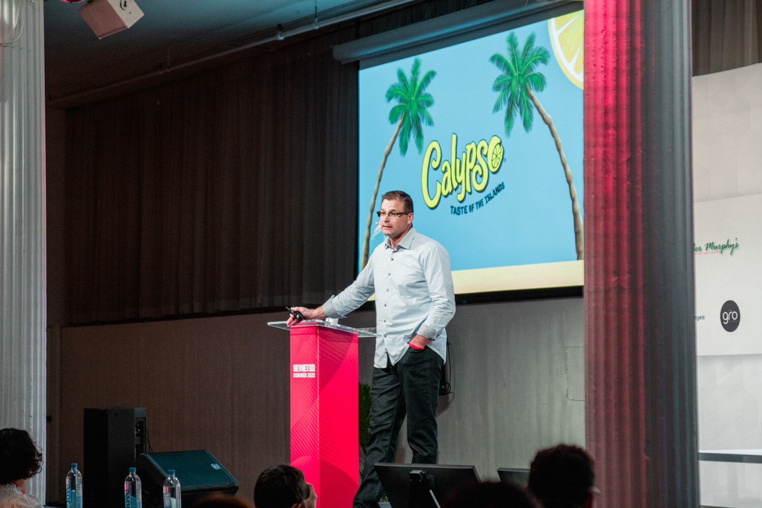 A man in a light blue shirt leans on a red podium in front of a blue slide with the Calypso Taste of the Islands logo and palm trees.