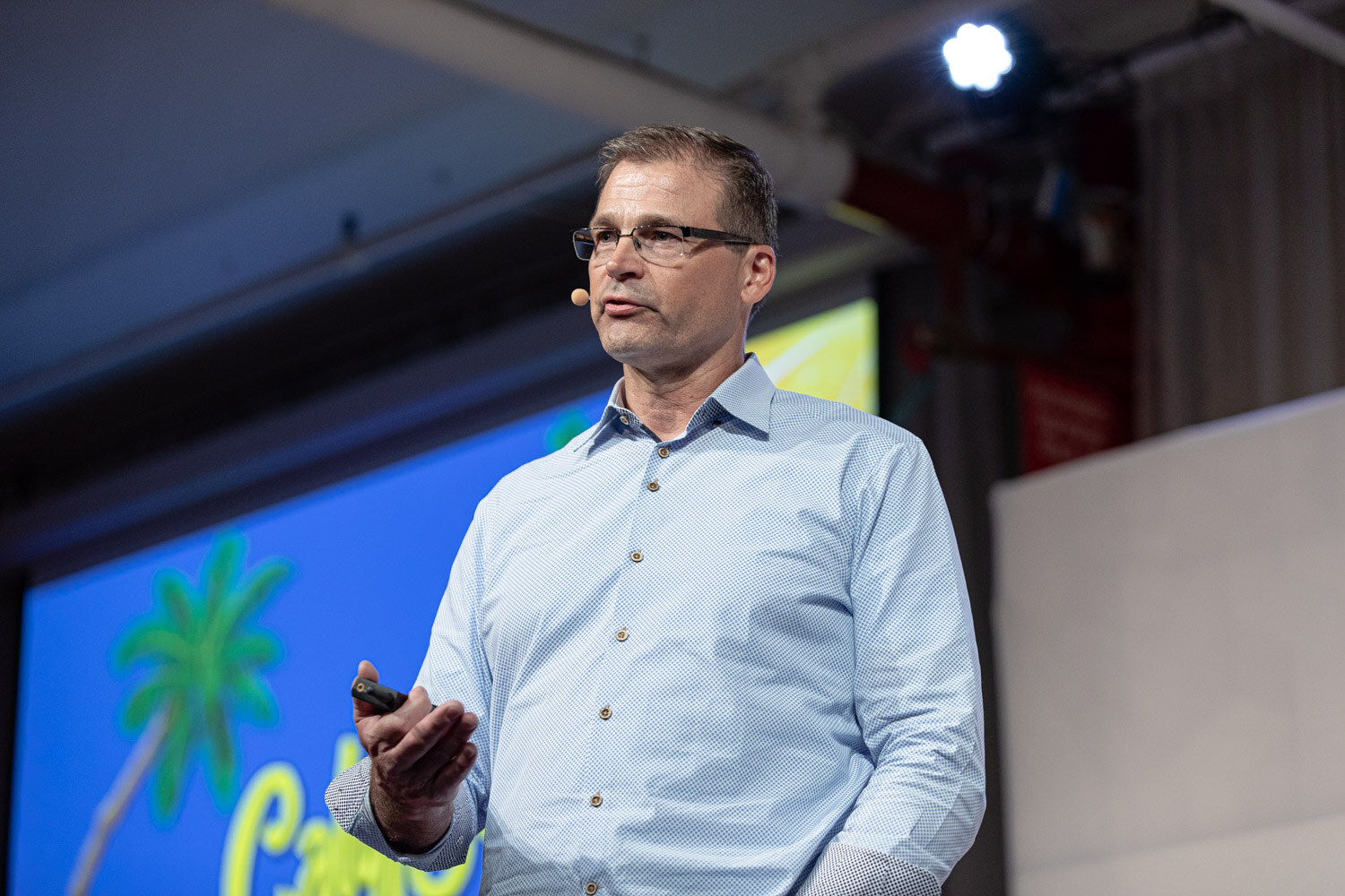 A man with glasses in a light blue shirt holds a clicker in front of a blue slide with a palm tree.