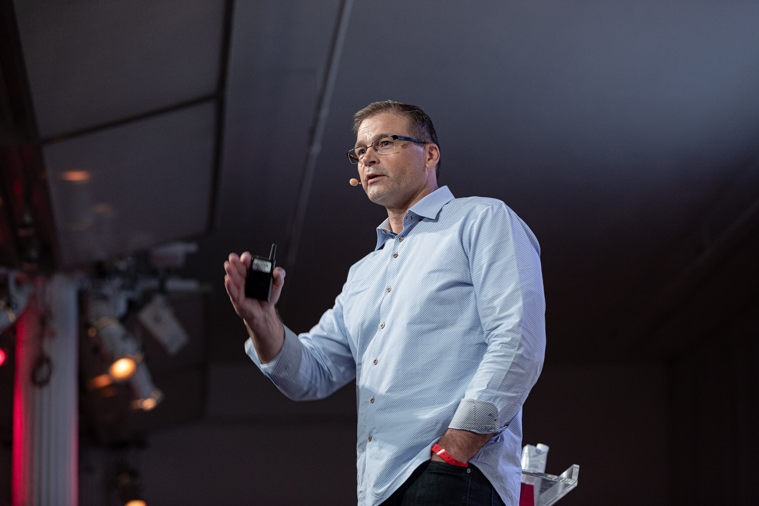 A man in a light blue shirt and glasses holds a clicker and raises one hand, the stage lights glowing behind him.