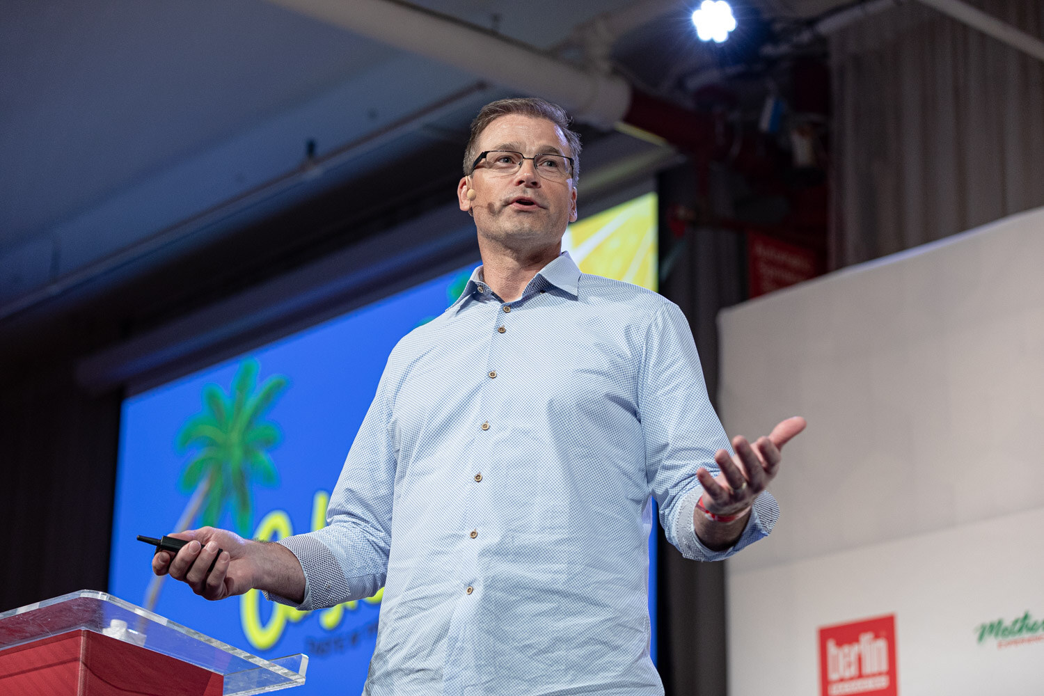 A man with glasses in a light blue shirt holds a clicker in front of a blue slide with a palm tree illustration.