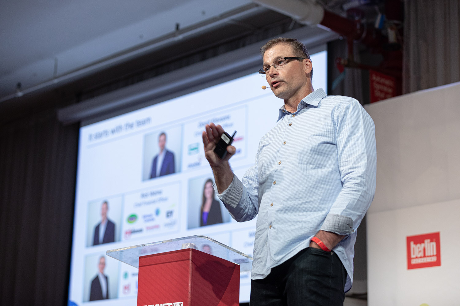 A man in a light blue shirt holds a clicker beside a podium as a slide reading It starts with the team shows photos of several people.