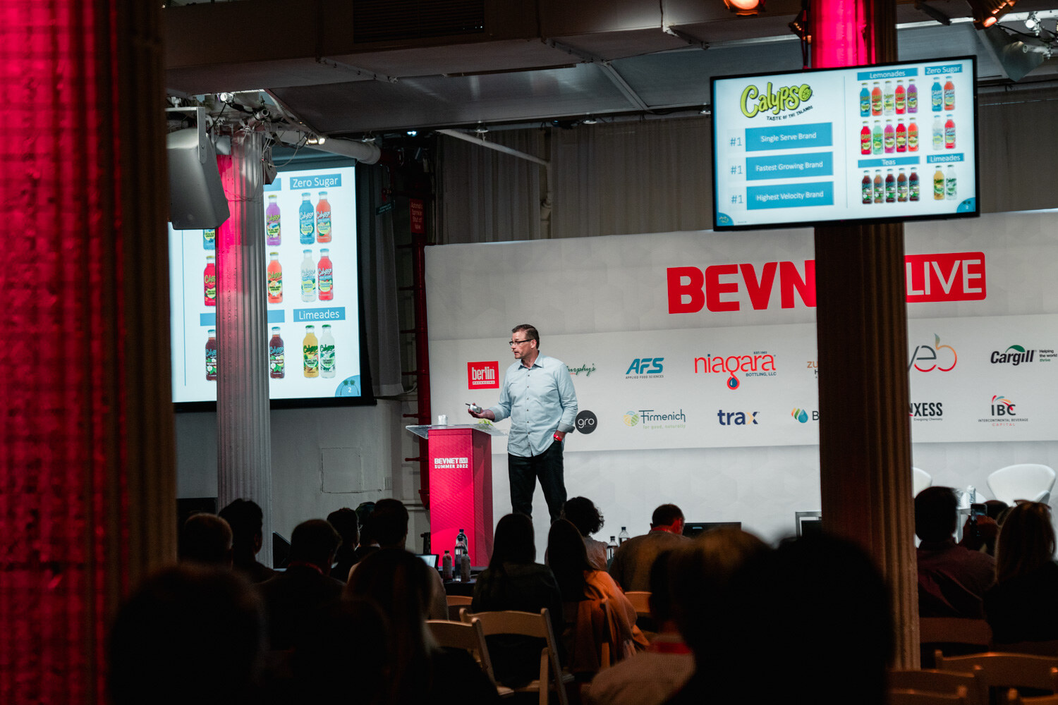 A man in a light blue shirt stands at a podium in a room with a red column, a monitor showing rows of bottles behind him.