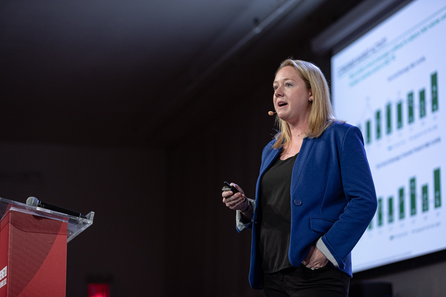A woman in a blue blazer holds a clicker with her other hand in her pocket in front of a slide of green bar charts.