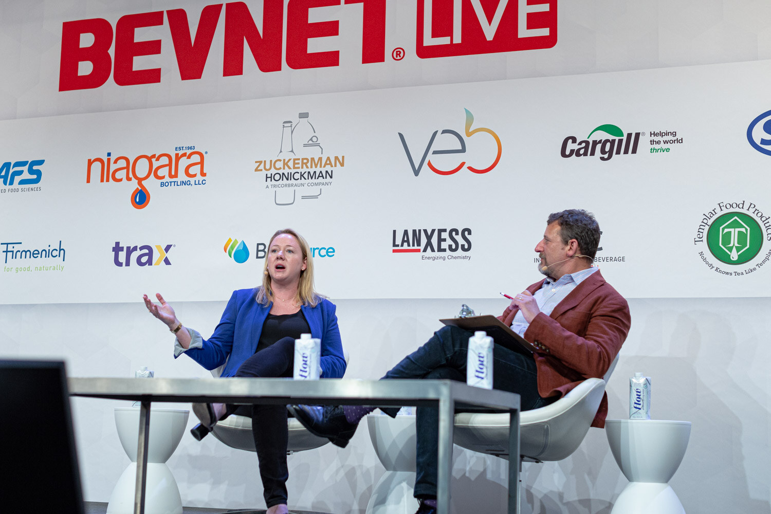 A woman in a blue blazer gestures with open hands while a man in a red jacket holds a tablet, both seated on stage.