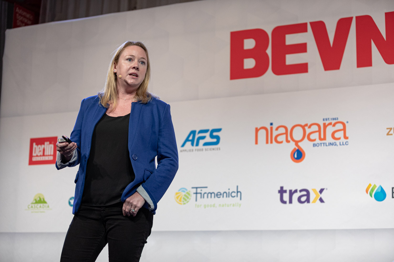 A woman in a blue blazer and black top stands holding a clicker, with Niagara, AFS and Trax logos behind her.