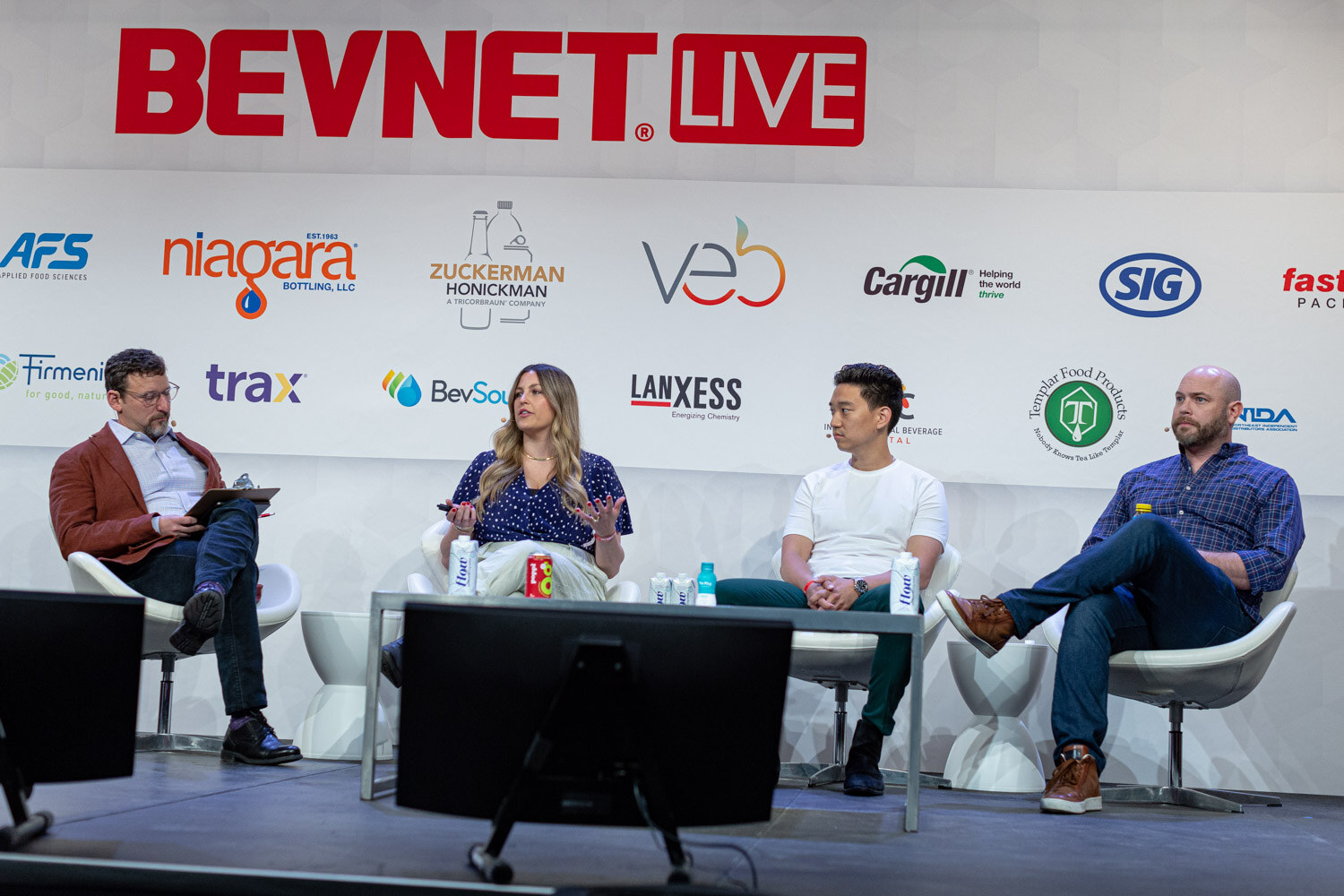 Four people seated in white chairs on the BevNET Live stage with a monitor and audience in the foreground.