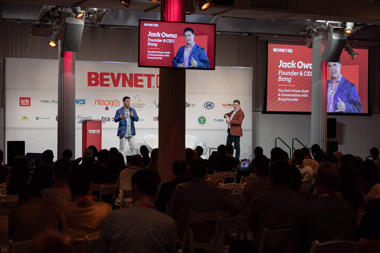 Wide view of a BevNET Live stage with two screens showing the Bang session slide, a speaker and an interviewer on stage, and the audience seated in front.
