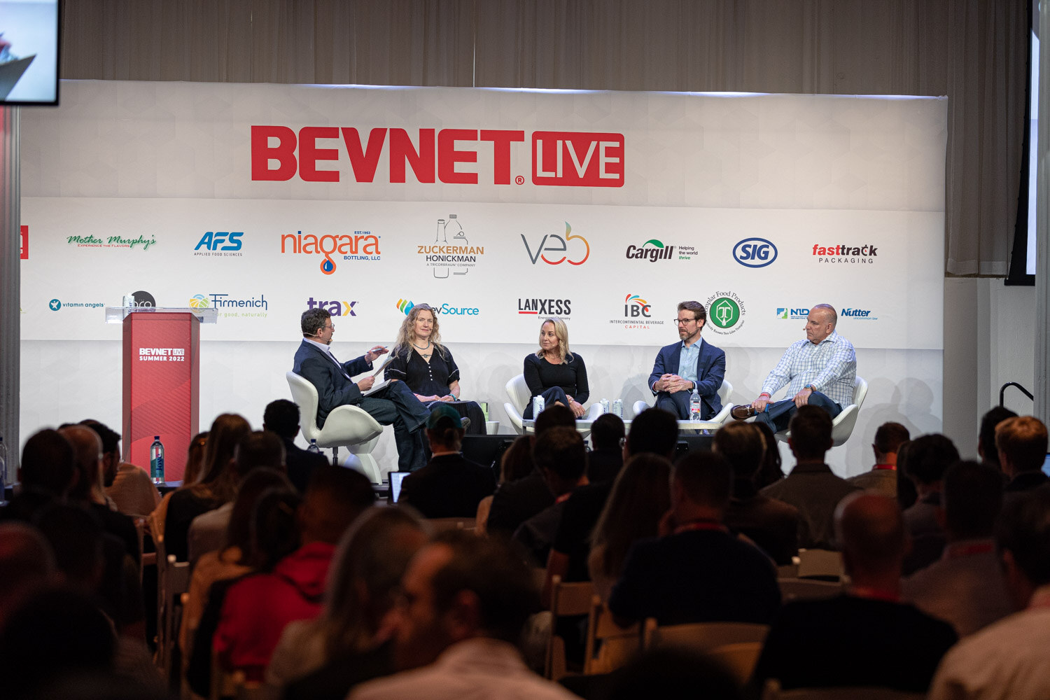 Five people seated in white chairs in front of a BevNET Live backdrop, with a crowd of attendees seen from behind.