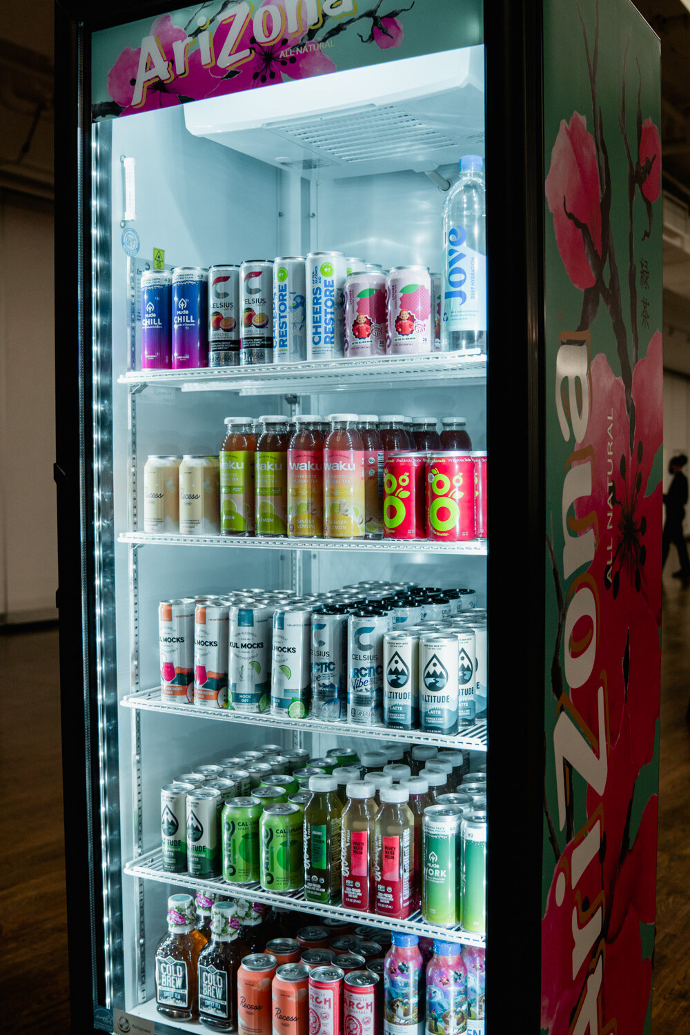 A tall glass-door refrigerator with an Arizona logo and pink blossom artwork, filled with canned and bottled drinks on lit shelves.