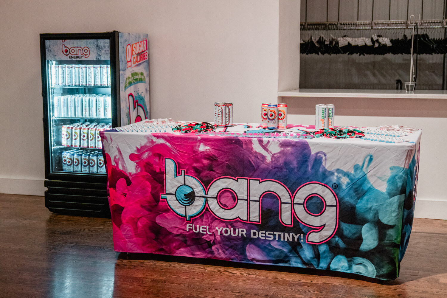 A table draped in a pink and blue Bang Fuel Your Destiny cloth with energy drink cans on top, next to a glass-door cooler of Bang cans.