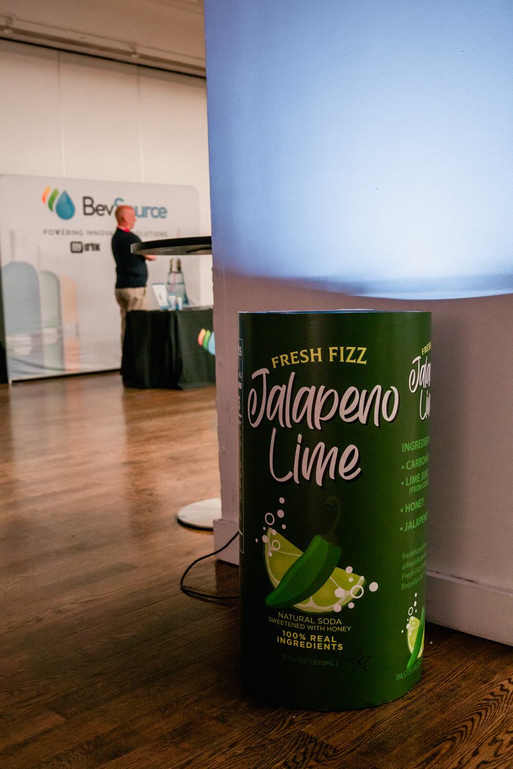 A green cylinder labeled Fresh Fizz Jalapeno Lime on a wood floor with a BevSource booth blurred in the background.