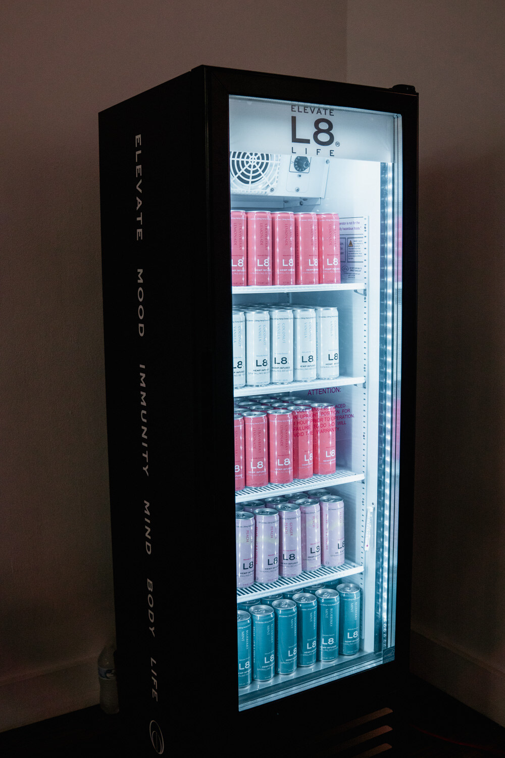 A black glass-door cooler labeled L8 Elevate Mood Immunity Mind Body Life with rows of cans, in a dim corner.