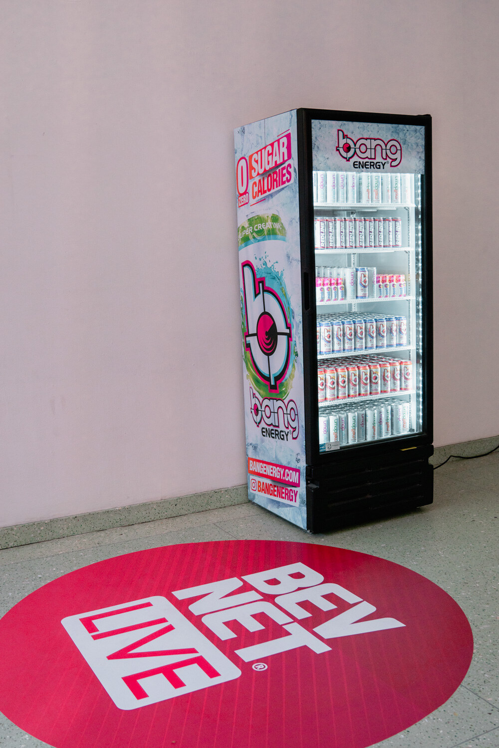 A red circular floor decal with the BevNET Live logo in front of a glass-door cooler with Bang Energy graphics.