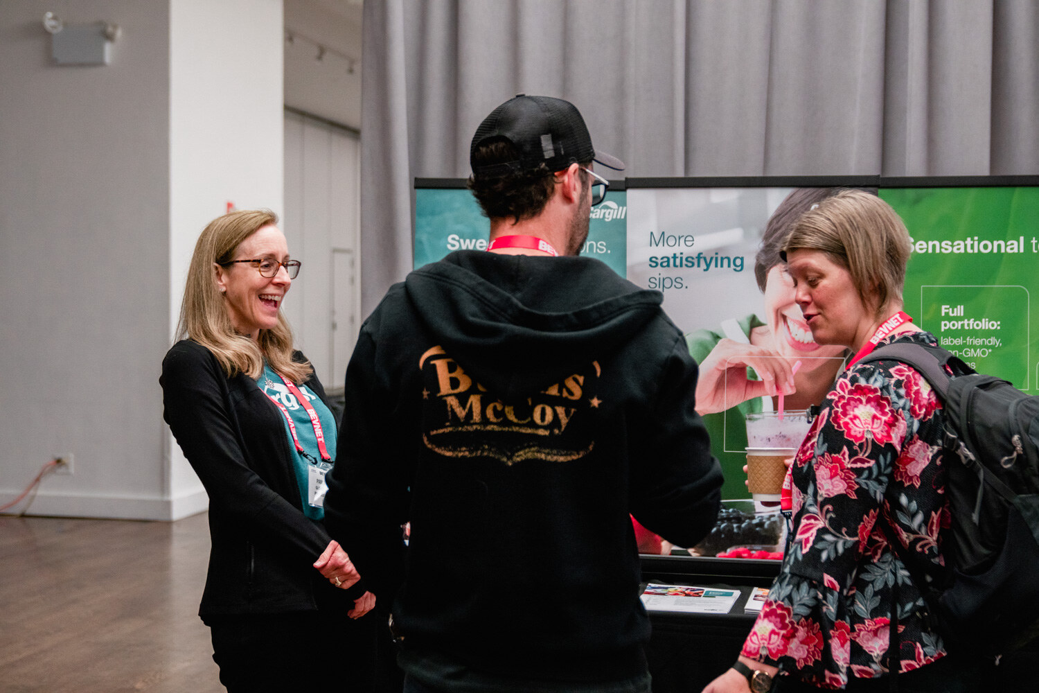 A woman with glasses laughs while a man in a black hoodie and a woman in a floral top talk in front of a green display banner.