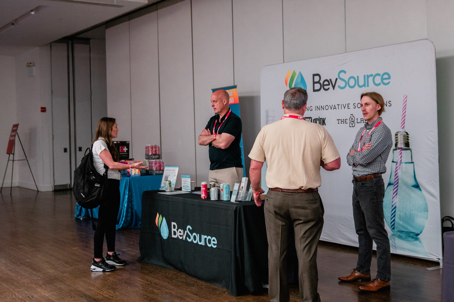 Several people stand around a table with a black BevSource tablecloth in front of a tall BevSource banner with a light bulb and straw graphic.