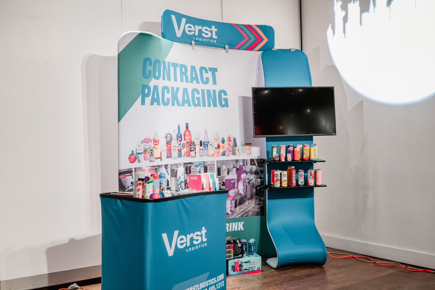 A blue and white booth reading Verst Logistics Contract Packaging with a screen and shelves of packaged drinks.