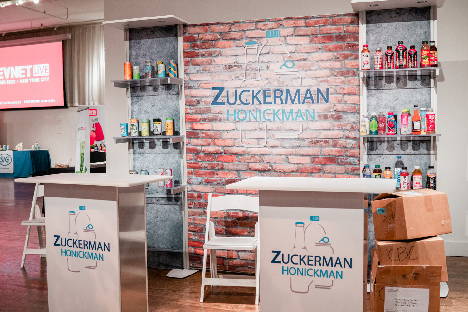 A booth with a faux brick wall reading Zuckerman Honickman, with shelves of bottles and cans and two white standing tables.