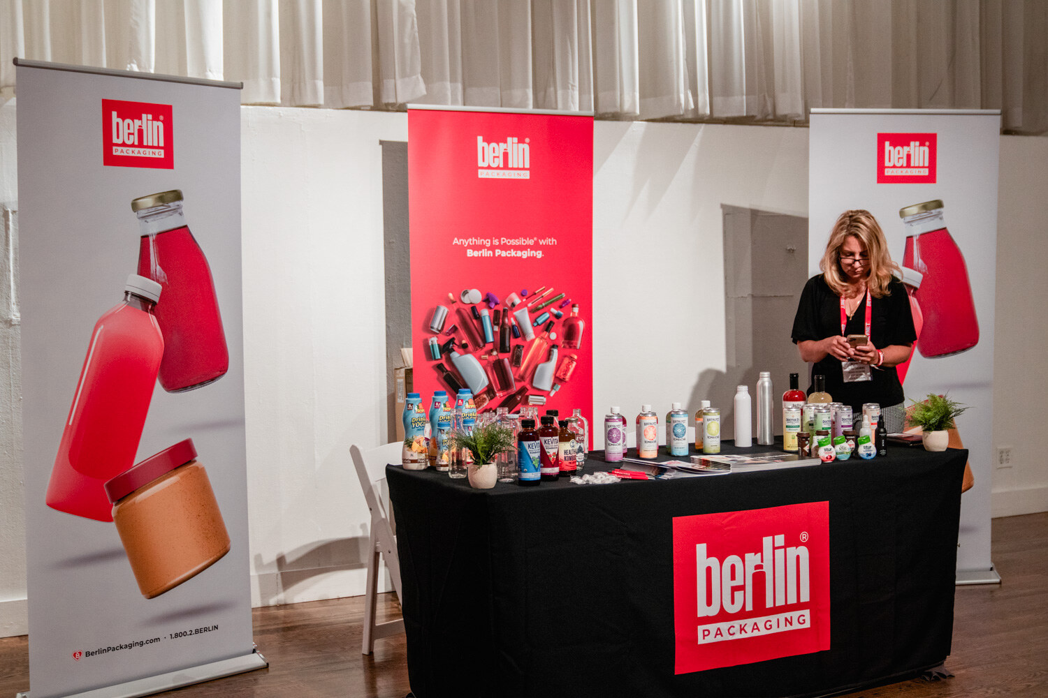 A woman in black stands behind a Berlin Packaging table with bottles and jars, with tall red banners showing bottles on both sides.