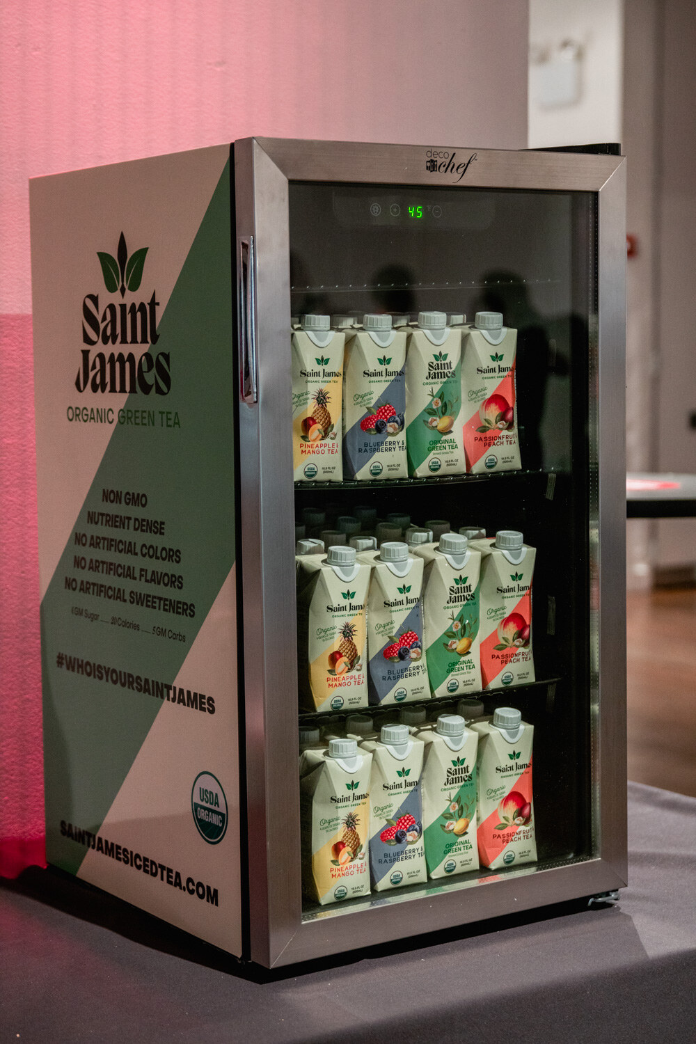 A small glass-front refrigerator stacked with cartons of Saint James Organic Green Tea with green and white graphics.