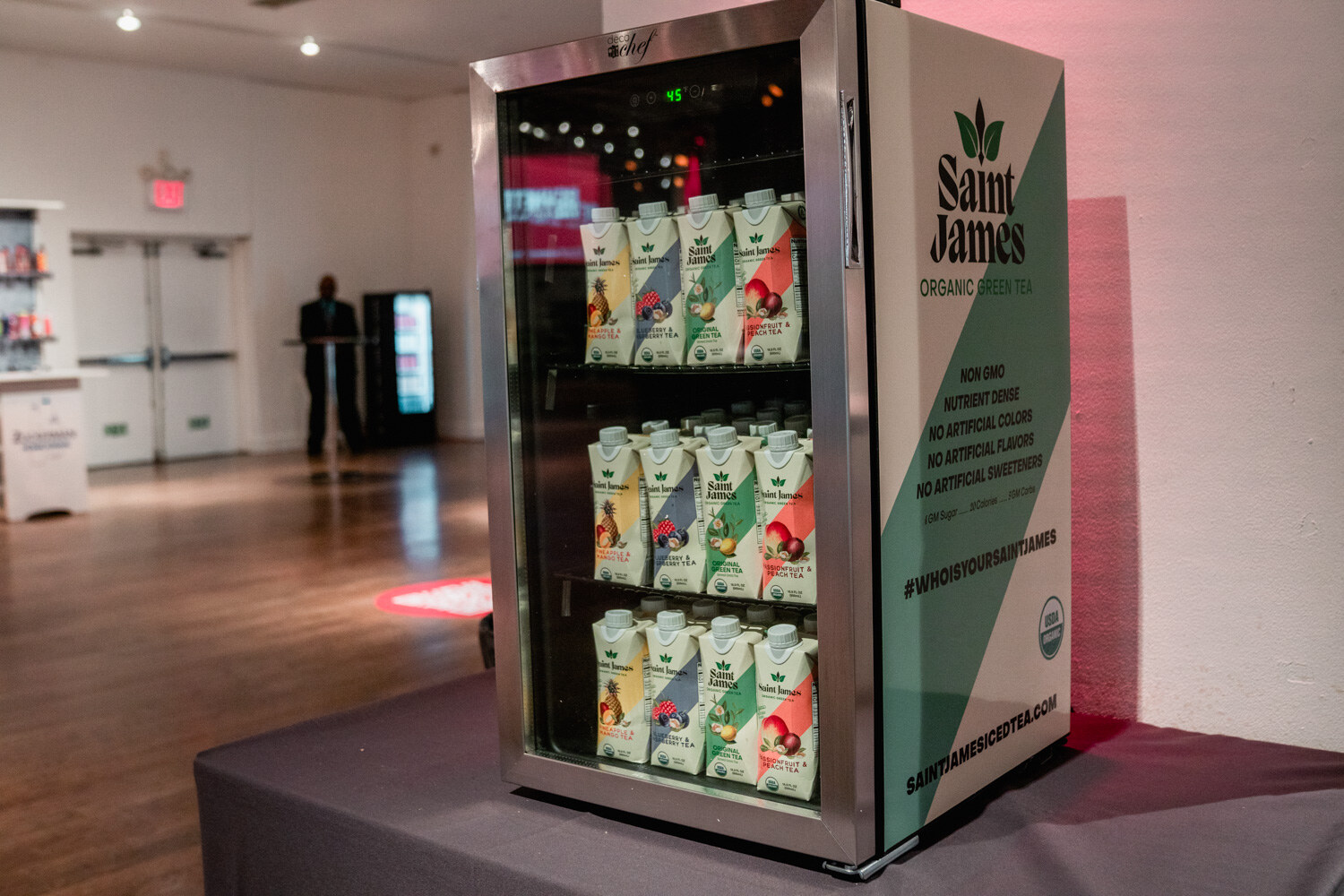 A small glass-door cooler filled with Saint James Organic Green Tea cartons with green and white graphics, on a platform in an open room.