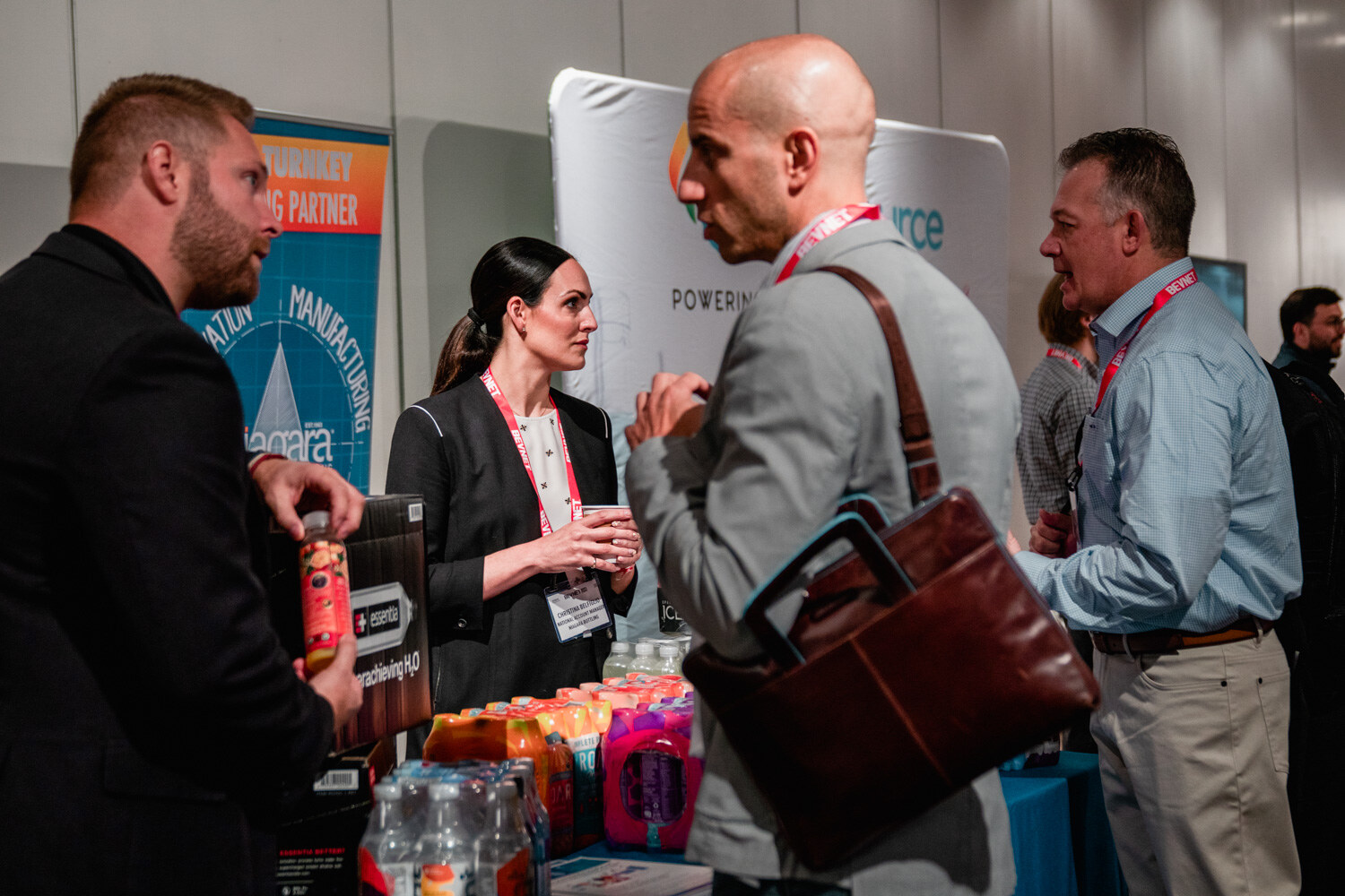 A woman in a black blazer talks to a bald man with a leather bag while others stand nearby, bottles of colored drinks on the table.
