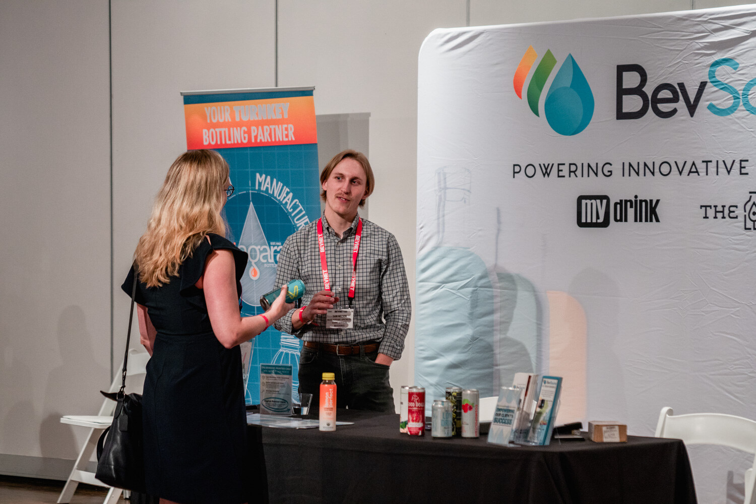 A man in a plaid shirt and a woman in a black dress talk at a table with cans, in front of a BevSource banner.