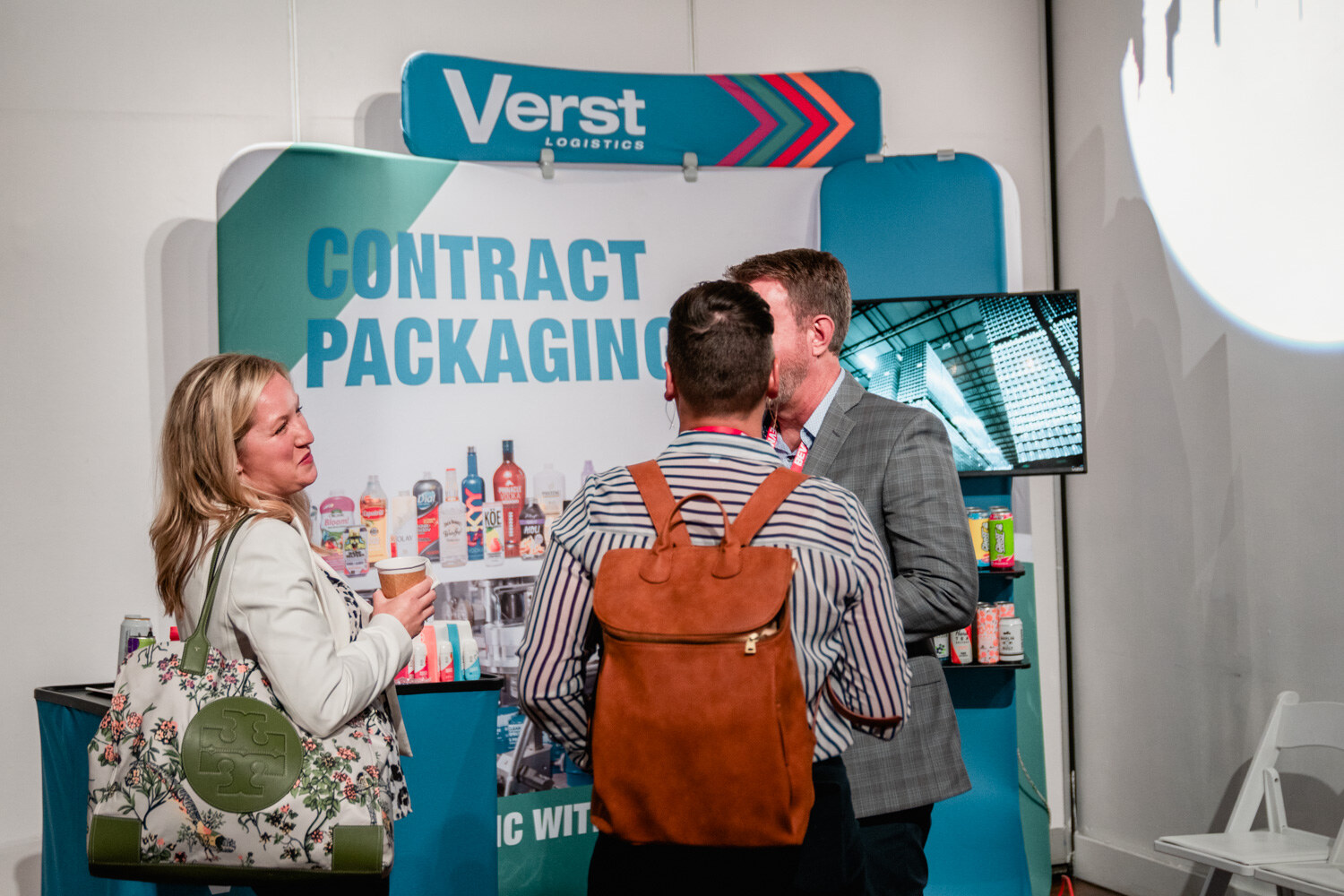 A woman with a floral tote and a man with a brown backpack talk with a man in a gray blazer in front of a Verst booth.